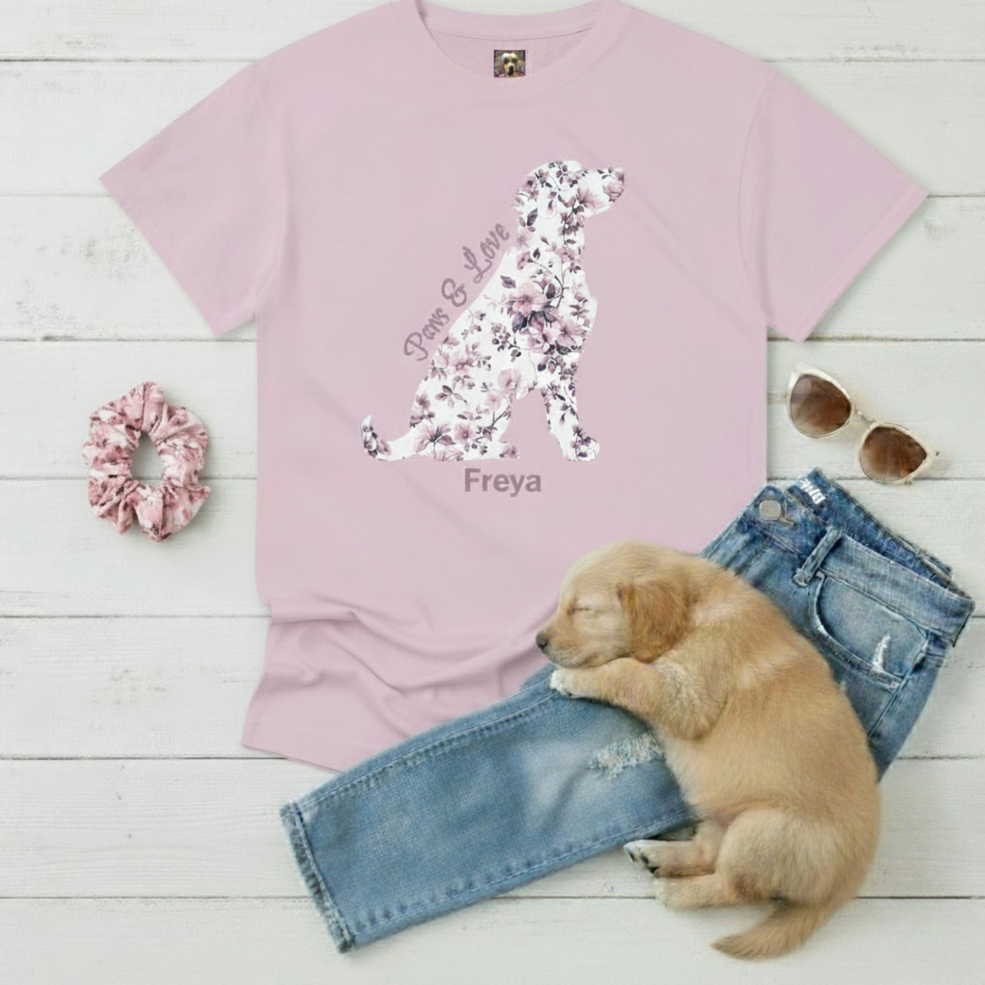 Pink t-shirt with pink French floral dog design, scrunchie, sunglasses, puppy dog, and jeans on a wooden surface