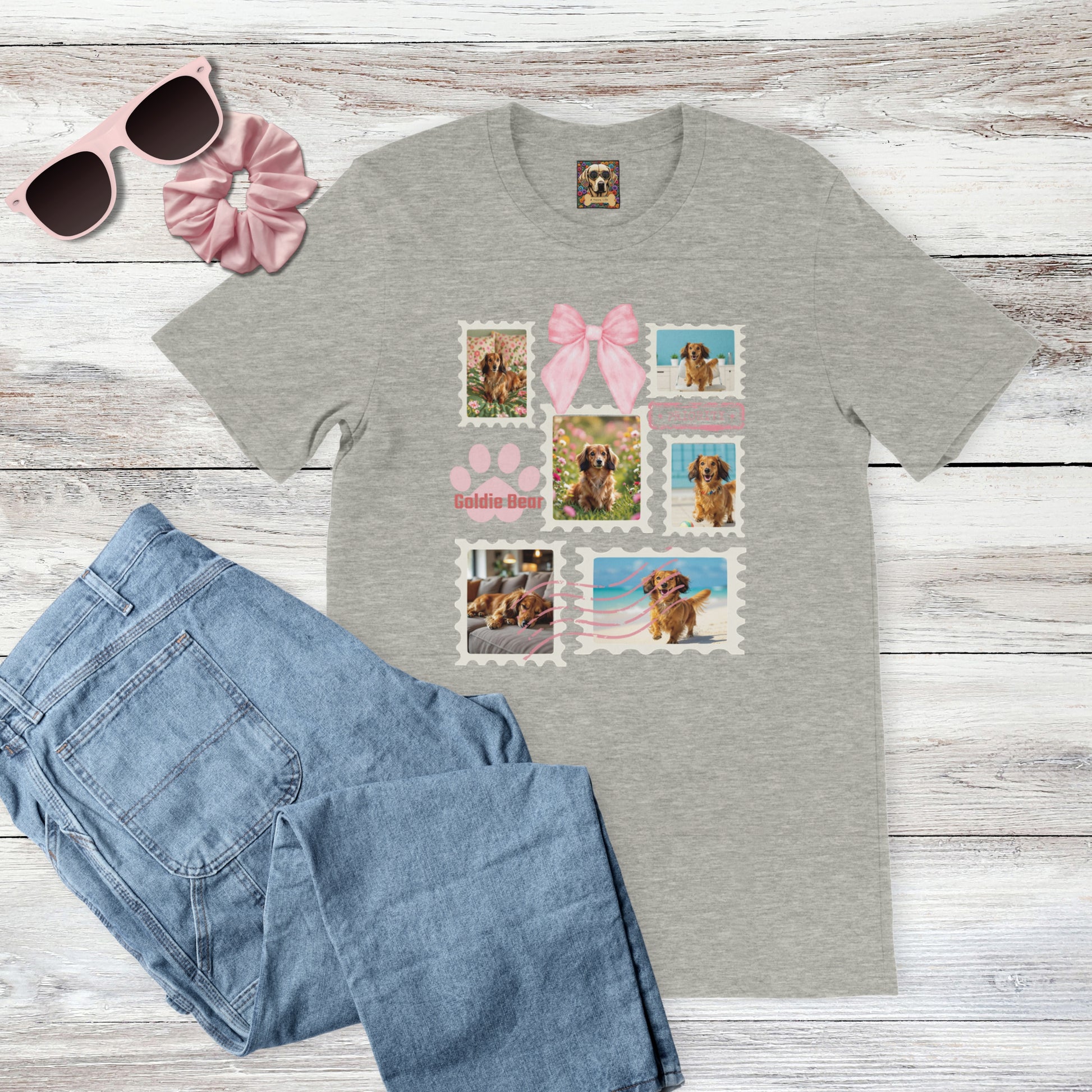 Personalized pet photo gray t-shirt with  design of a postage stamp collage and pink bow next to jeans, pink sunglasses, and hair scrunchie on a wooden surface