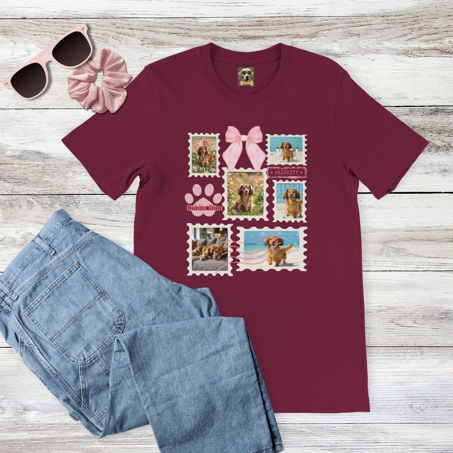 Personalized pet photo maroon t-shirt with  design of a postage stamp collage and pink bow next to jeans, pink sunglasses, and hair scrunchie on a wooden surface