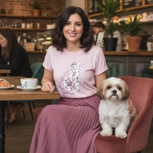 Woman in a light pink graphic t- shirt that has a french pink floral in the shape of a Shih Tzu on it and with a dog sitting  next to her in a cafe.