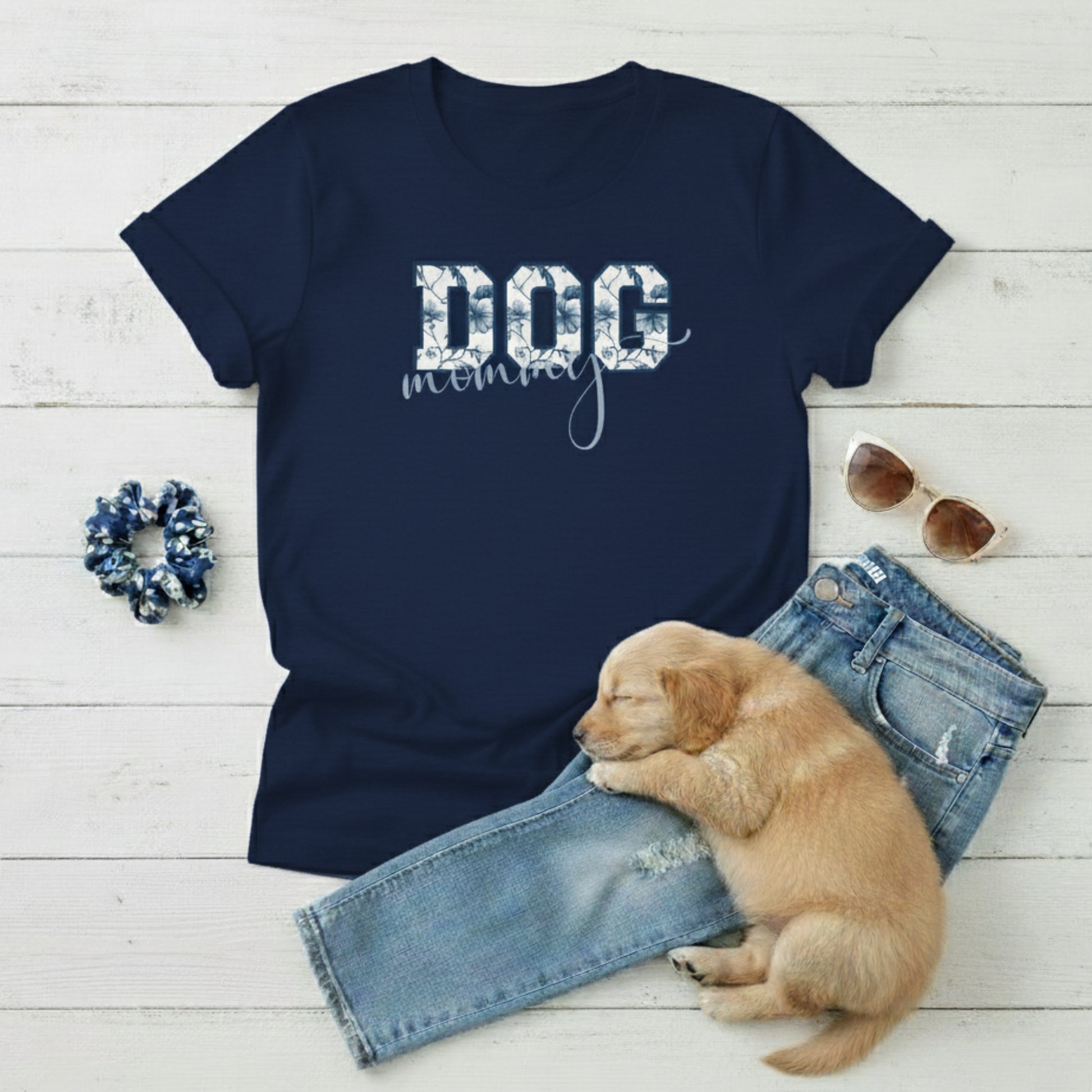 Navy blue Dog Mom t-shirt with 'DOG' text in Toile De Jouy print and mommy in script font. A cute puppy, scrunchie, sunglasses, and jeans on a light wooden surface suggest how to style this fitted tee.