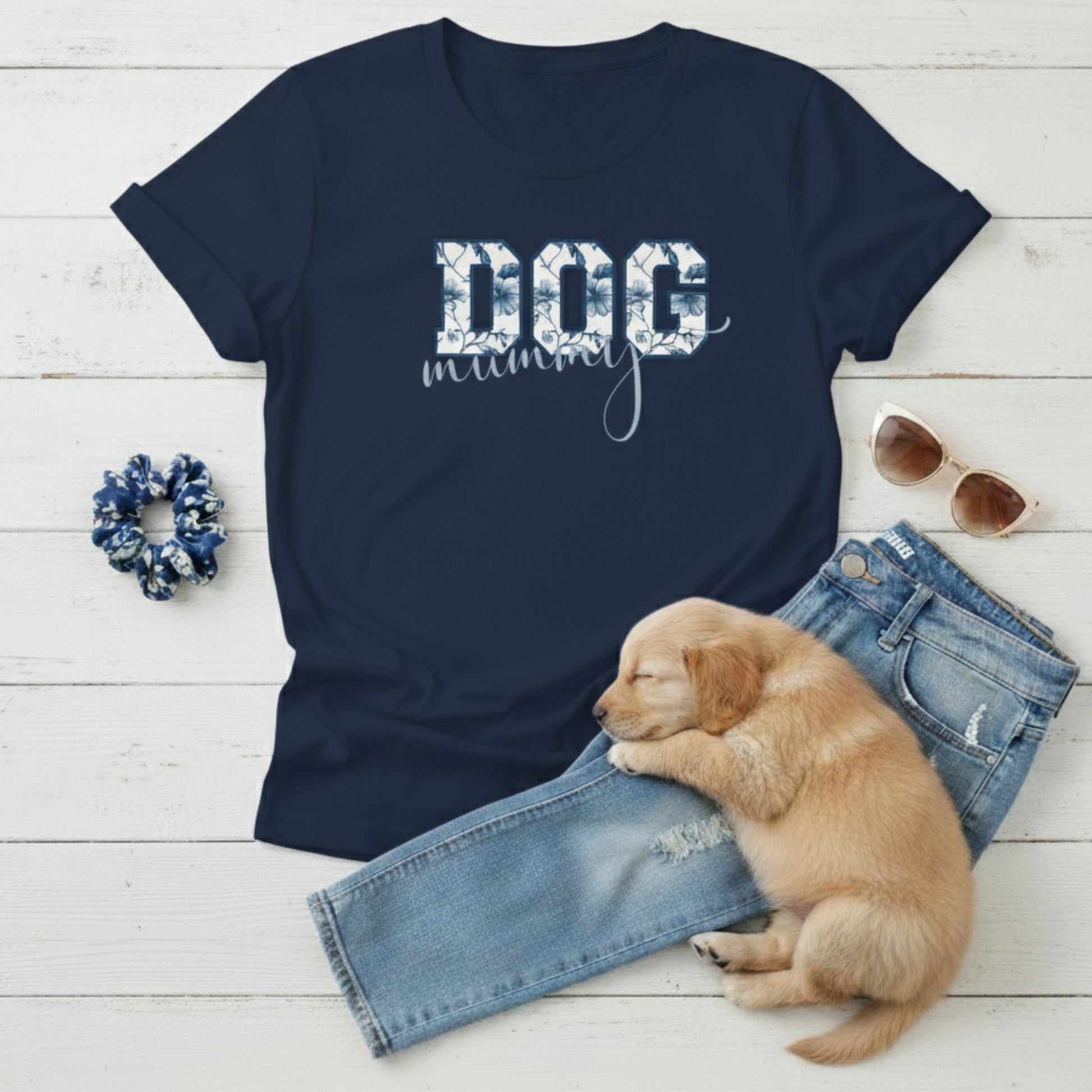 Navy blue Dog Mom t-shirt with 'DOG' text in Toile De Jouy print and mummy in script font. A cute puppy, blue scrunchie, cat eye retro sunglasses, and faded denim jeans on a light wooden surface suggest how to style this fitted tee.