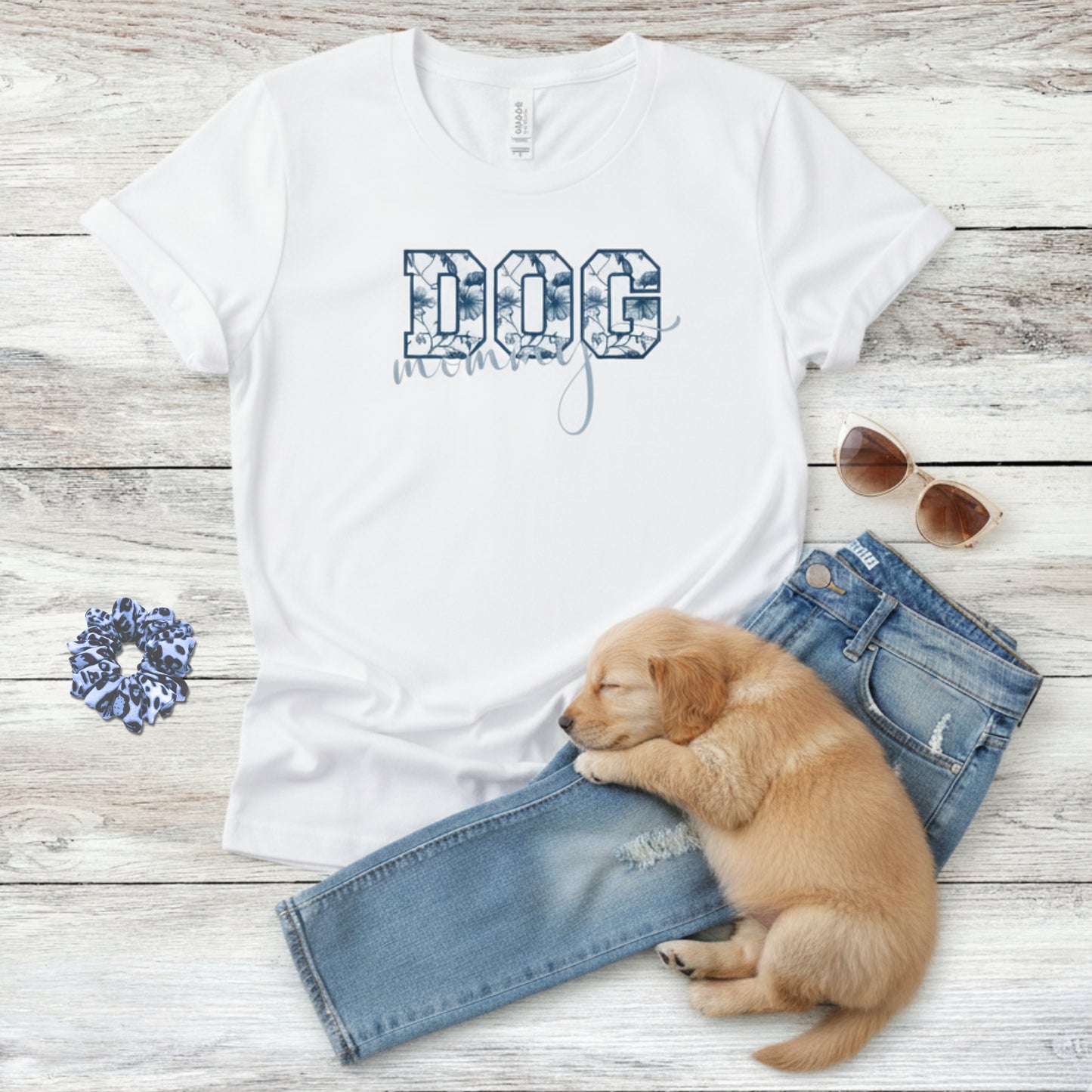 Bella Canvas 6004 white Dog Mom t-shirt with bold 'DOG' text in blue floral, Toile De Jouy, print and mommy in script font. A cute sleeping puppy, blue scrunchie, cat eye retro sunglasses, and faded denim jeans on a light wooden surface suggest how to style this fitted tee.