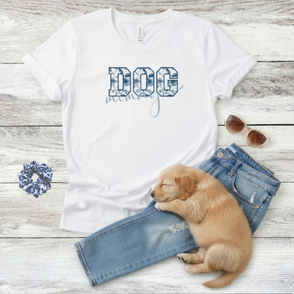Bella Canvas 6004 white Dog Mom t-shirt with bold 'DOG' text in blue floral, Toile De Jouy, print and mommy in script font. A cute sleeping puppy, blue scrunchie, cat eye retro sunglasses, and faded denim jeans on a light wooden surface suggest how to style this fitted tee.