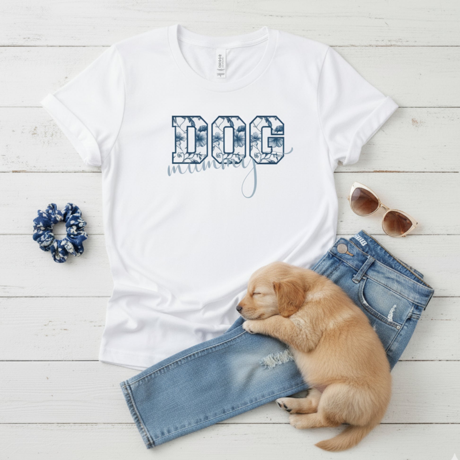 Bella Canvas 6004 white Dog Mom t-shirt with bold 'DOG' text in blue floral, Toile De Jouy, print and mummy in script font. A cute sleeping puppy, blue scrunchie, cat eye retro sunglasses, and faded denim jeans on a light wooden surface suggest how to style this fitted tee.