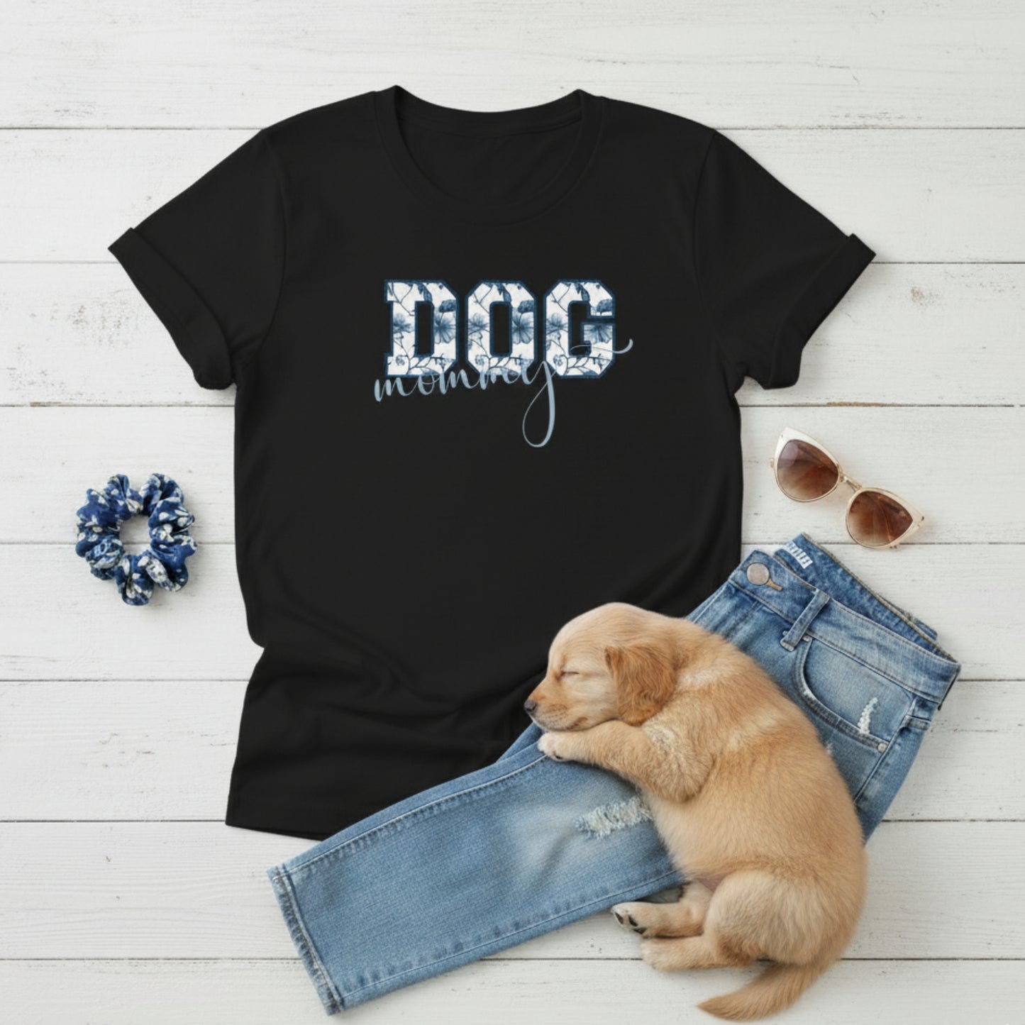 Bella Canvas 6004 black Dog Mom t-shirt with bold 'DOG' text in blue floral, Toile De Jouy, print and mommy in script font. A cute puppy, blue scrunchie, cat eye retro sunglasses, and faded denim jeans on a light wooden surface suggest how to style this fitted tee.
