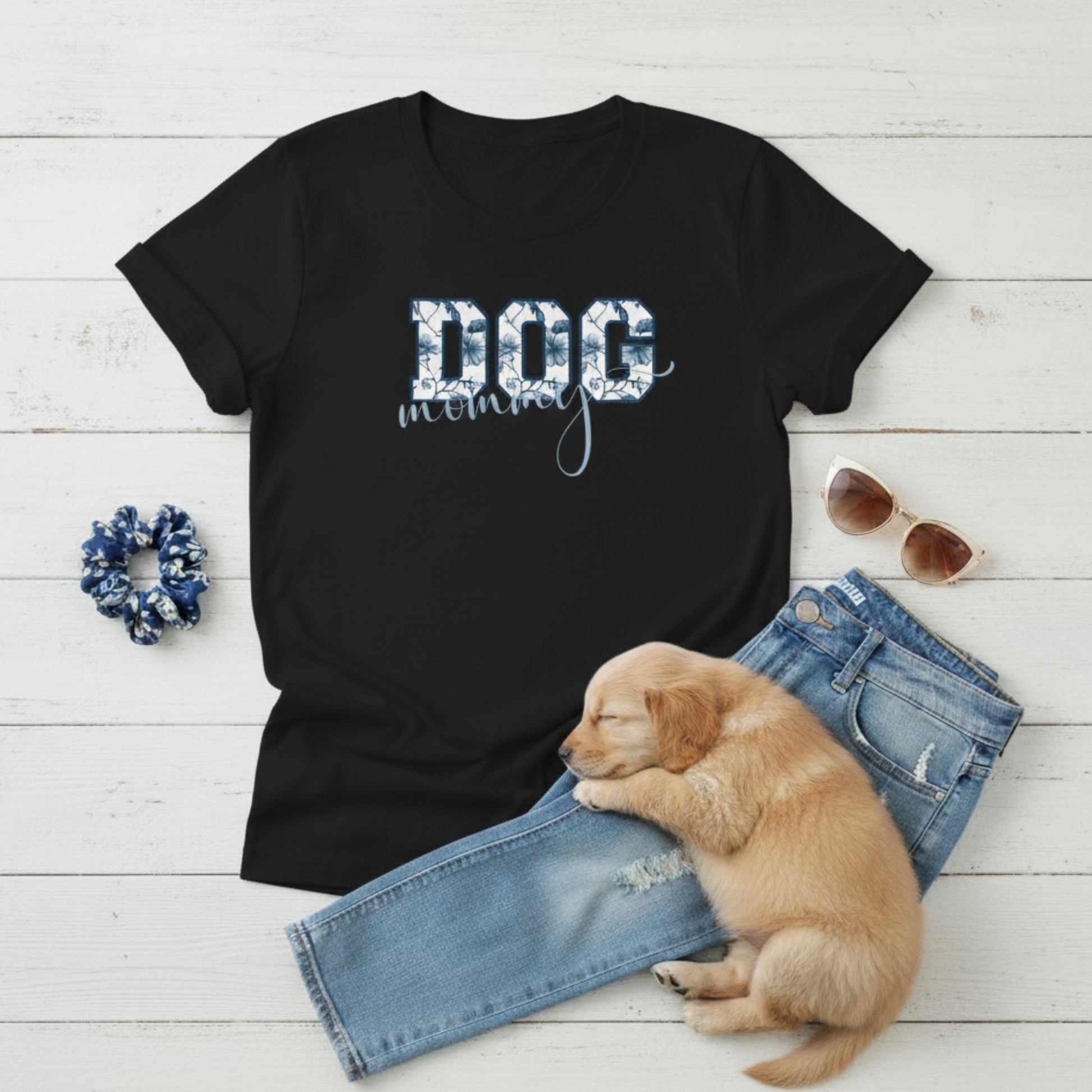 Bella Canvas 6004 black Dog Mom t-shirt with bold 'DOG' text in blue floral, Toile De Jouy, print and mommy in script font. A cute puppy, blue scrunchie, cat eye retro sunglasses, and faded denim jeans on a light wooden surface suggest how to style this fitted tee.