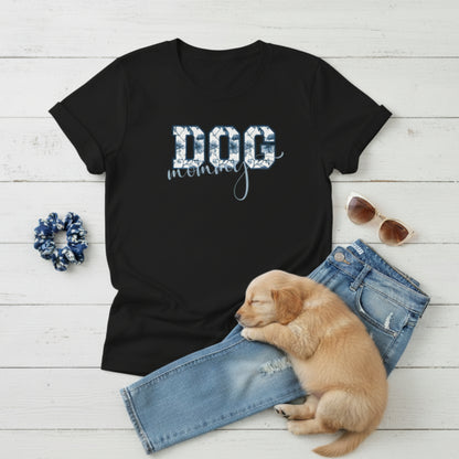 Bella Canvas 6004 black Dog Mom t-shirt with bold 'DOG' text in blue floral, Toile De Jouy, print and mommy in script font. A cute puppy, blue scrunchie, cat eye retro sunglasses, and faded denim jeans on a light wooden surface suggest how to style this fitted tee.