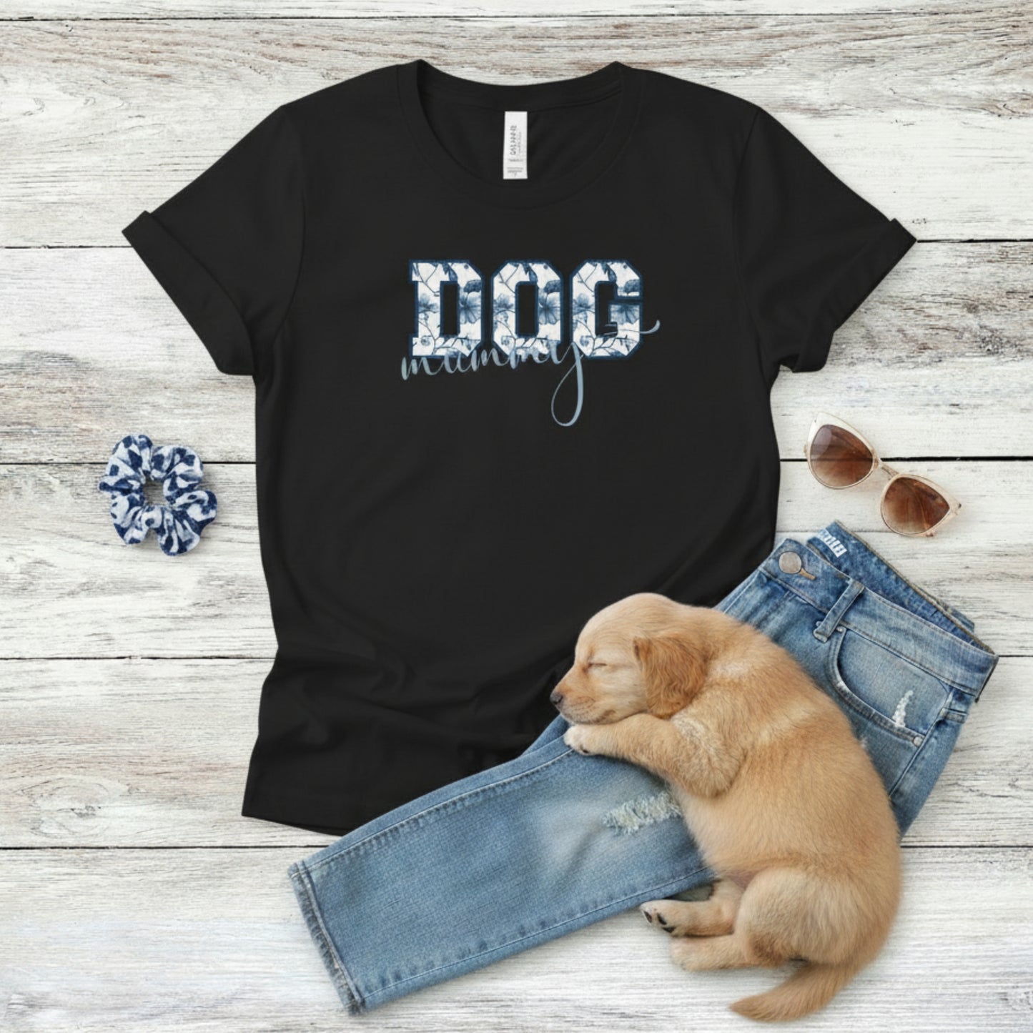 Bella Canvas 6004 black Dog Mom t-shirt with bold 'DOG' text in blue floral, Toile De Jouy, print and mummy in script font. A cute puppy, blue scrunchie, cat eye retro sunglasses, and faded denim jeans on a light wooden surface suggest how to style this fitted tee.