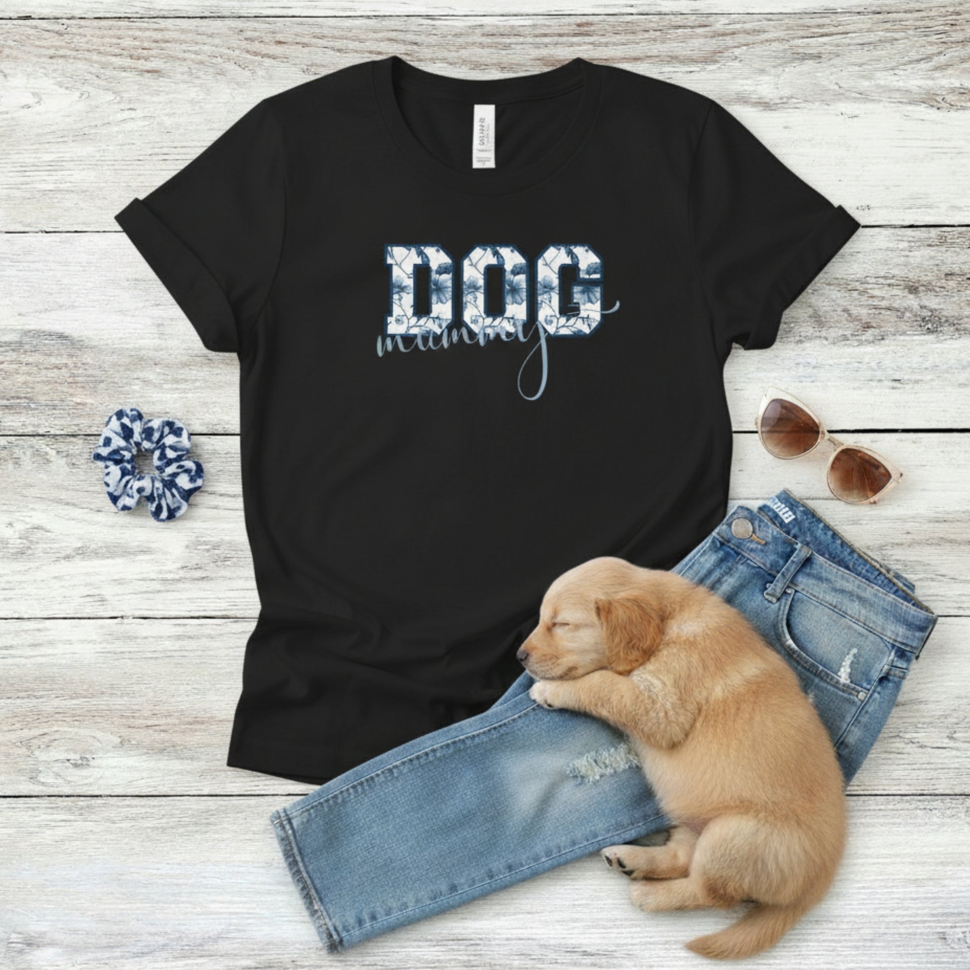 Bella Canvas 6004 black Dog Mom t-shirt with bold 'DOG' text in blue floral, Toile De Jouy, print and mummy in script font. A cute puppy, blue scrunchie, cat eye retro sunglasses, and faded denim jeans on a light wooden surface suggest how to style this fitted tee.