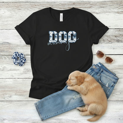 Bella Canvas 6004 black Dog Mom t-shirt with bold 'DOG' text in blue floral, Toile De Jouy, print and mummy in script font. A cute puppy, blue scrunchie, cat eye retro sunglasses, and faded denim jeans on a light wooden surface suggest how to style this fitted tee.
